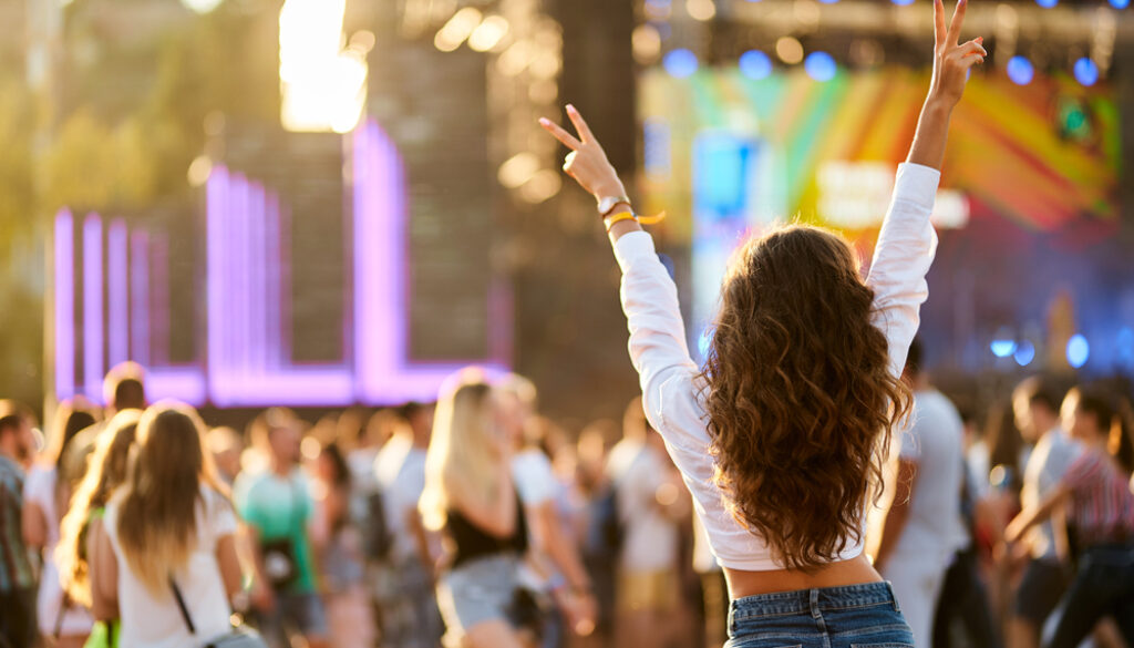 Back view of joyful woman with raised hands enjoys summer music festival. Crowd dances at beach concert, sunset light. Happy fan cheers at outdoor live event. Excited attendee celebrates, vibes.