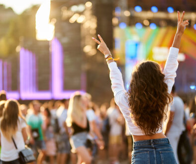Back view of joyful woman with raised hands enjoys summer music festival. Crowd dances at beach concert, sunset light. Happy fan cheers at outdoor live event. Excited attendee celebrates, vibes.